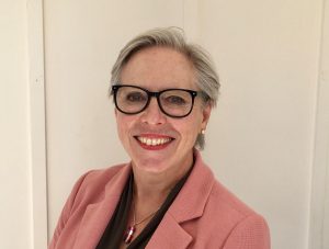 Photo of Auriol who is wearing a pink blazer, with a black top and has black rimmed glasses, with a short pixie cut and grey hair