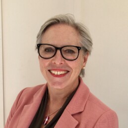 Photo of Auriol who is wearing a pink blazer, with a black top and has black rimmed glasses, with a short pixie cut and grey hair